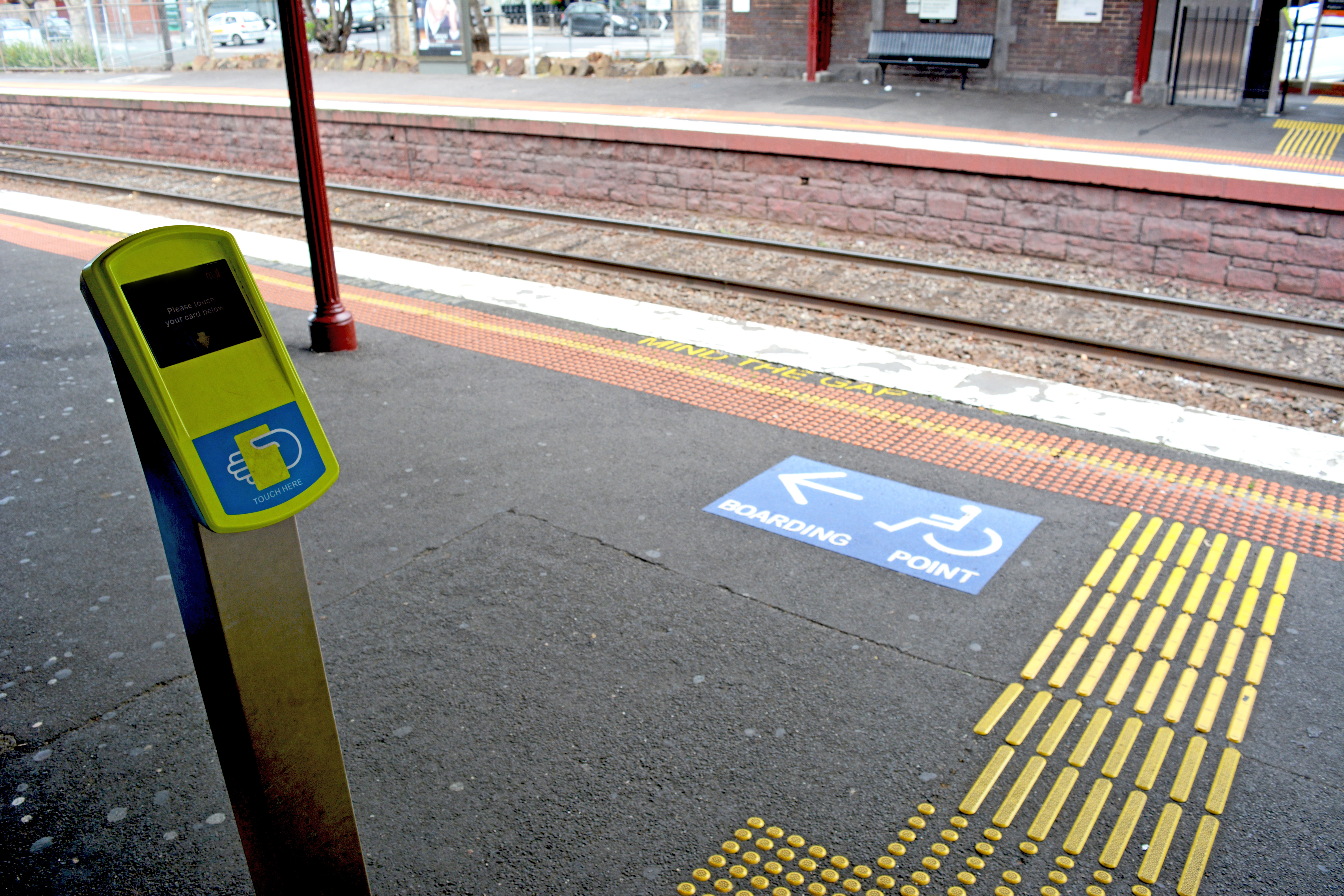 Photo of a platform with a directional boarding point decal and a myki machine. The words “Mind the Gap” have been stencilled onto the platform next to the edge. 