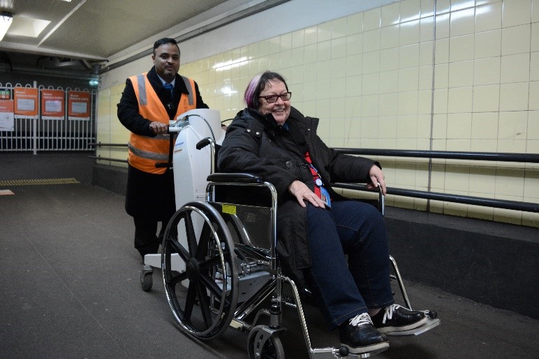 Image of a smiling station staff member using a Wheelchair Mover to assist a passenger up a steep ramp. The passenger is also smiling.